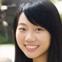 Young Asian woman with long black hair smiling at the camera, outdoor blurred greenery in background.